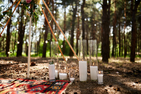 Candles burning in glass vases, flasks on carpet near bohemian tipi arch decorated with roses and flowers wrapped in fairy lights on outdoor wedding ceremony venue in pine forest. Boho rustic decor.