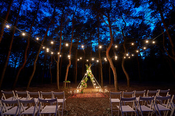 Festive string lights illumination on boho tipi arch decor on outdoor wedding ceremony venue in pine forest at night. Vintage string lights bulb garlands shining above chairs at summer rural wedding. © artiemedvedev