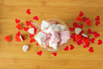 Top view of glass double walls cup of coffee with marshmallow. On the wooden table with red paper hearts and candies.