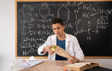 Young Arabic chemistry teacher holding a piece of hamburger. Student eating fast food in the...
