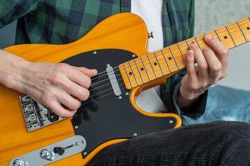 A man in a plaid shirt plays a natural-colored electric guitar sitting on a bed in close-up, selective focus.A male musician plays an electric guitar.Telecaster of natural color.Person playing guitar.