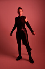 Full length portrait of pretty redhead female model wearing black futuristic scifi leather cloak costume. Standing pose  holding lightsaber on red studio background with coloured lighting
