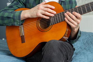 A man in a plaid shirt plays a natural-colored classical guitar sitting on a bed in close-up, selective focus. A male musician plays the guitar. Classical guitar in natural color.