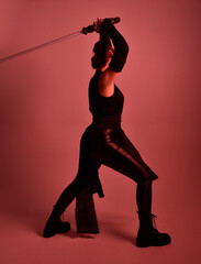 Full length portrait of pretty redhead female model wearing black futuristic scifi leather cloak costume. Standing pose  holding lightsaber on red studio background with coloured lighting