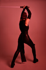 Full length portrait of pretty redhead female model wearing black futuristic scifi leather cloak costume. Standing pose  holding lightsaber on red studio background with coloured lighting