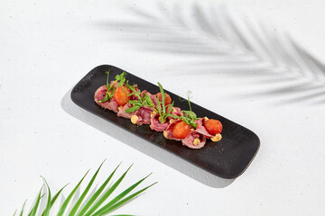 Roast beef with cherry tomatoes, ruccola and spicy sauce on black plate. Sliced roast beef on white concrete background. Italian antipasti in summer style with hard shadow and palm leaves.