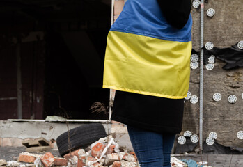 Obraz premium Woman with Ukrainian flag looking at abandoned destroyed building. Russia invasion, war in Ukraine, crisis and destruction concept. Girl standing in front of ruined, bombed residential house.