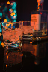A bar counter with two glasses of strong white alcohol with ice. Next to the glasses is a cigar, and against the background is a bottle of strong drink and the blurred lights of the bar illumination.