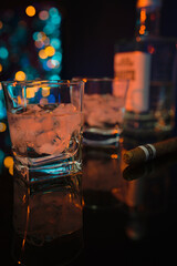 Two glasses with ice and strong alcohol on the bar counter. A cigar and a bottle of strong white alcohol on the bar. In a blurry background, colored lights of the bar illumination.