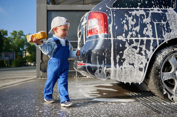 Kid boy studying work at car wash. Toddler child, holding yellow foam sponge in his right hand,...