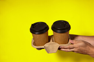 Close-up on a yellow background, women's hands hold two paper cups with coffee in a stand. Coffee or tea with you. Promotion two for the price of one, coffee delivery on a yellow background
