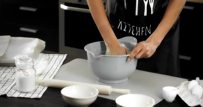Woman in a brown apron is standing at a table in a modern kitchen, kneading dough with her hands in a gray bowl to bake shortbread cookies. Baking Ingredients