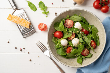 Fresh salad with arugula, cherry tomatoes, mozzarella cheese and hard cheese on white wooden background. Top view