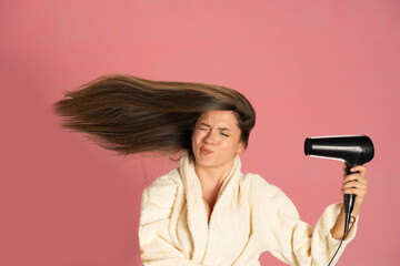 Funny woman drying her long hair with electric fan on a pink background - obrazy, fototapety, plakaty