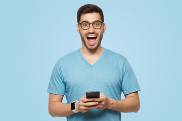 Young handsome man holding phone in hands, looking at camera with excited WOW face