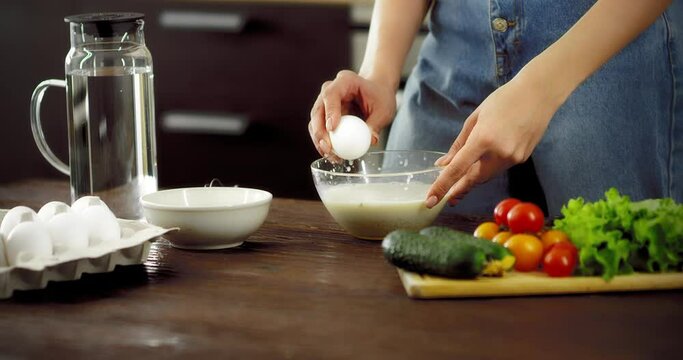 Young woman in jeans cooks breakfast in a modern kitchen, breaks eggs by injuring the shell into a bowl. Concept of a healthy lifestyle