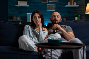 POV of focused girlfriend and boyfriend watching movie on television, eating popcorn and drinking beer from bottle. Young couple doing fun activity with film on TV channel program.