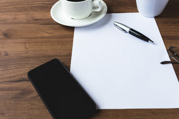 An office desk with a cup of coffee, a mobile phone, glasses, a pen and a white sheet of paper.Top view mock-up
