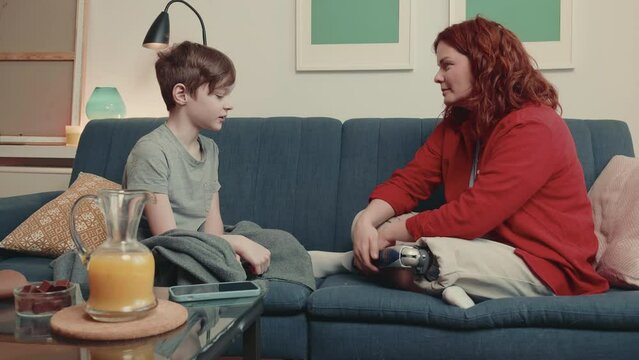 Happy young Caucasian teen age boy touches womans bionic leg prosthesis sitting together on couch at cozy home.