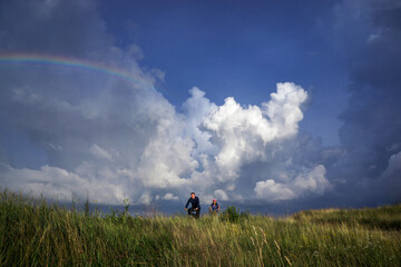 A man and a woman ride bicycles in a field under a high blue cloudy sky with a rainbow.