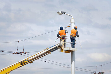 Painter team in lift bucket paint street lighting pole at height. Men in cradle painting lamppost with paint brush. Cherry picker with workers at height, risky job. Renovate street lightning pole © Tricky Shark