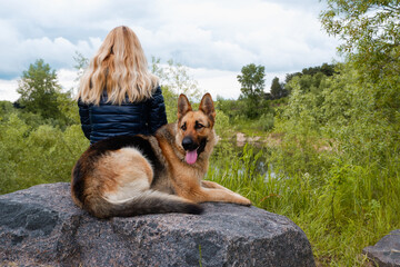 A young woman with a German Shepherd sits on a large granite stone by the lake on a cloudy spring day. Young woman relaxing with her friend dog at the coast sitting on a stone promenade looking out © Trik