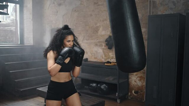 Strong woman kicks punching bag with boxing gloves in gym
