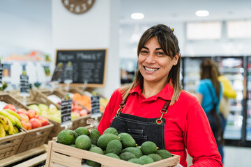 Latin woman working in supermarket holding a box containing fresh avocados - 499099052