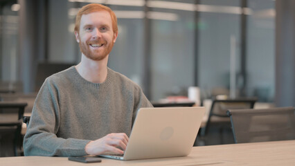 Young Man Smiling at Camera while using Laptop in Office