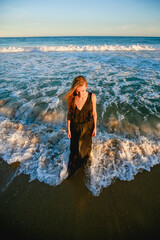 A woman with blond hair blowing in the wind is standing in the sea in a long evening dress. Evening sunset light.