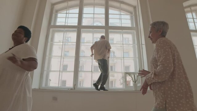 Dolly out shot of Caucasian senior man moonwalking on windowsill while his diverse female friends dancing energetically at bright spacious dance studio