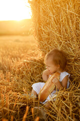 A hungry child eats a loaf sitting by a large haystack. Delicious sweet pastries. A girl in nature at sunset. The sun's rays. A hot summer day