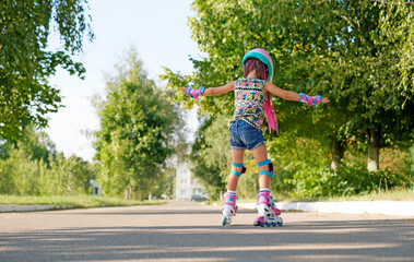 Kid tries to keep his balance and not fall, for the first time standing on roller skates. view behind. A girl is learning to rollerblade. Active sports for summer recreation and outdoor entertainment