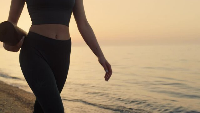 Fit girl holding mat walking seashore at sunset. Sportswoman going to training.