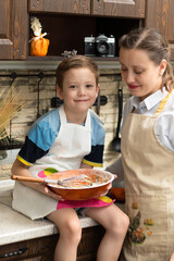 Beautiful young mom cook in an apron with her son prepares cookie dough in a wooden bowl at home in the kitchen. Selective focus. Portrait. Close-up