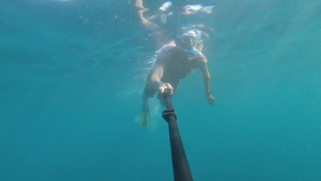 A Caucasian man in a snorkel mask and flippers dives and swims underwater with an action camera on a long selfie stick. snorkeling freediving