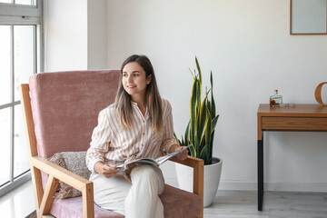 Young woman with magazine sitting in comfortable armchair at home