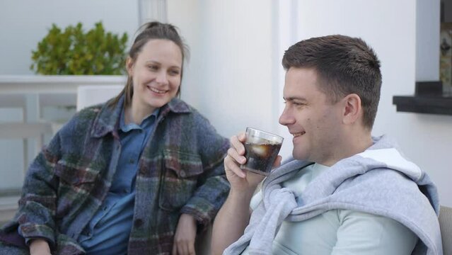 Causacian couple at the cafe terrace. Young man laughs sipping carbonated soft drink from the glass with ice and lemon slice.