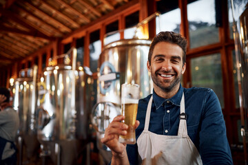 What total bliss. Portrait of a cheerful young business owner holding up a glass of beer that he just poured inside of a beer brewery during the day.