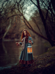 Magical girl with lantern in her hand standing near the river in the warm colored park