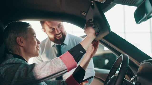 Close-up of happy brunette female testing buying car from inside during visit auto dealership. Female in car showroom talking to sales person and looking at automotive vehicle in dealership.