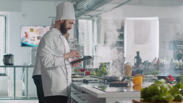 Male cook analyzing culinary recipe on digital tablet in kitchen, using internet on gadget to prepare food. Professional chef in uniform cooking gourmet meal from online gastronomy website.
