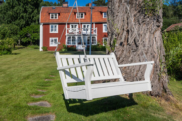 A white wooden garden swing hanging on a tree. Classical swinging bench on the green lawn and trees surrounded. Landscape design: beautiful flowers on foreground. Red traditional cabin in background.