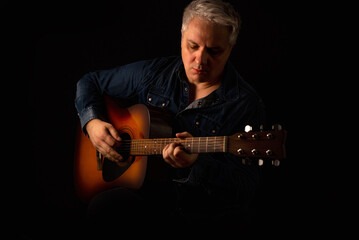 Session guitar player in a jeans shirt plays an acoustic guitar shot on black background. Low key portrait.