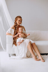 Fototapeta na wymiar Attractive redhead woman with long braidhair in white summer dress sitting on white sofa and holding her baby boy with her hands