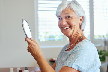 Still gorgeous after all these years. Portrait of a happy senior woman using a hand mirror at home.