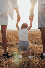 Close Up Back View of Mom and Dad Helping Their Child Making First Steps, Young Family Walking on the Field at Sunset