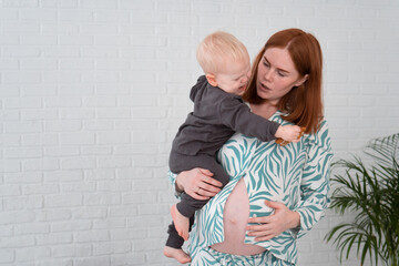 Worried redhead pregnant woman in pajamas holding little baby (crying boy) in her arms indoor