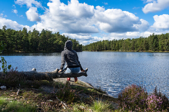 Alone beautiful girl on the forest coast of Scandinavia on fall sunny day. Young slim woman female traveler dressed in hoodie and blue jeans sitting relaxing watching panorama lake water in autumn.