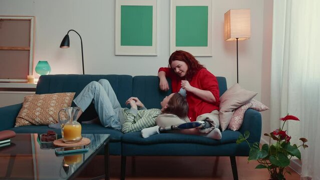 Happy beautiful serene 35-40 disabled mother with bionic prosthetic leg talking, bonding to teen age daughter on couch.